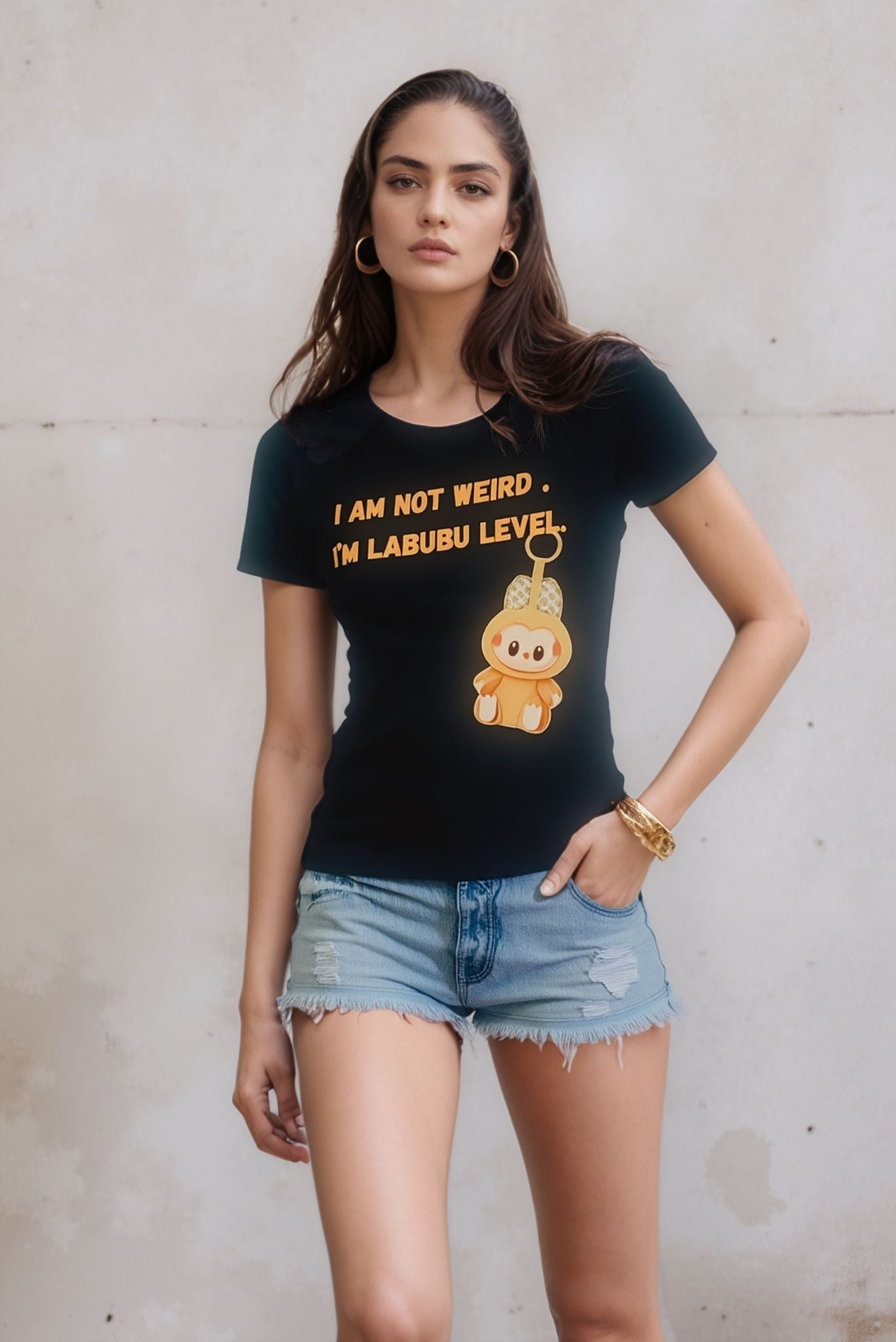 A woman wearing a black graphic t-shirt that says "I am not weird, I'm Labubu level" with a cute cartoon character illustration, paired with distressed denim shorts, standing against a plain light-colored wall.