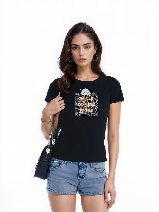 Woman wearing a black graphic t-shirt with the text "Smile. It Confuses People," paired with denim shorts and a black shoulder bag. She stands against a white background with a confident expression