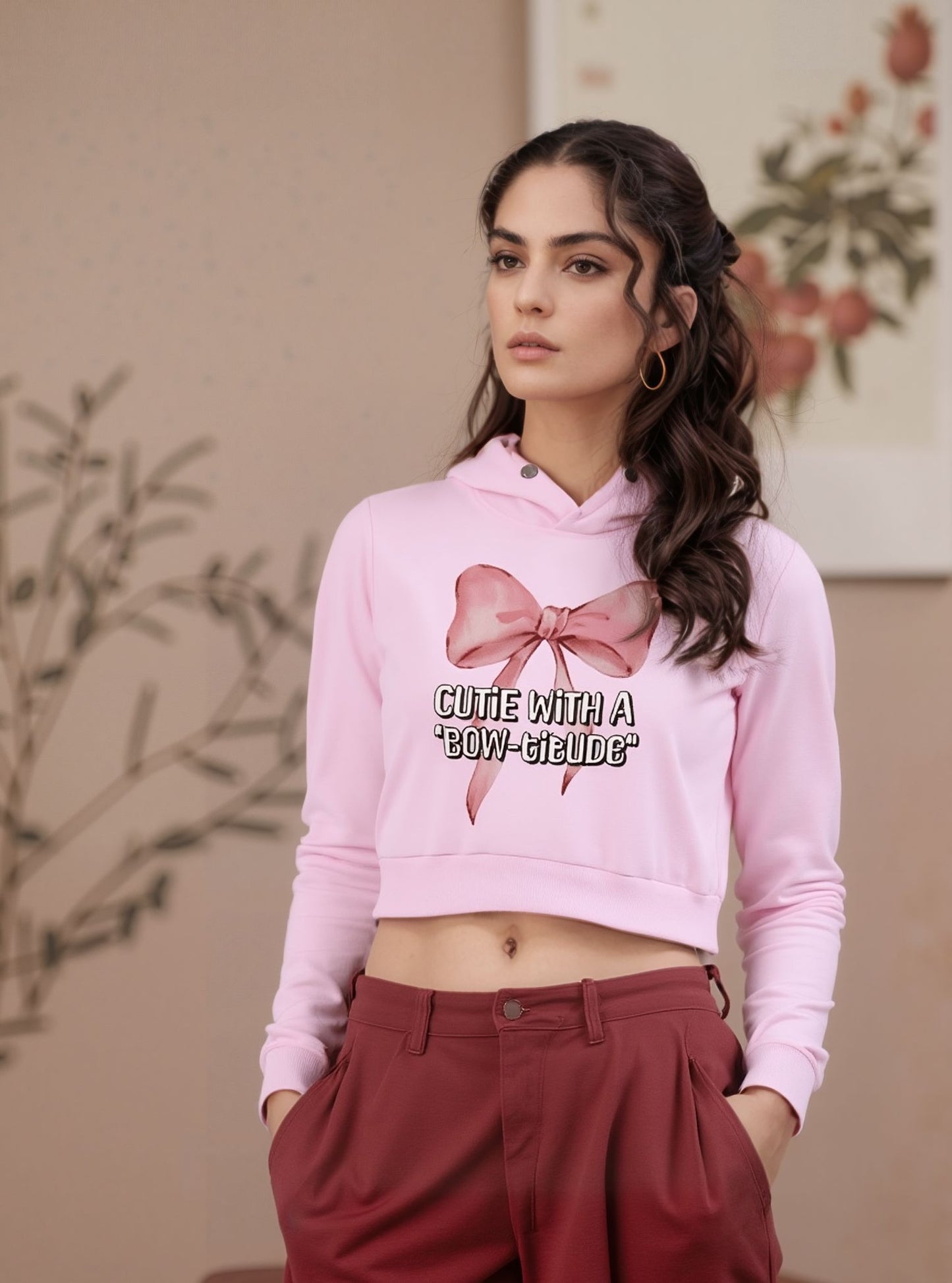 Woman posing in a pink cropped hoodie with a bow print and slogan “Cutie with a Bow-titude,” styled with high-waisted maroon trousers.

