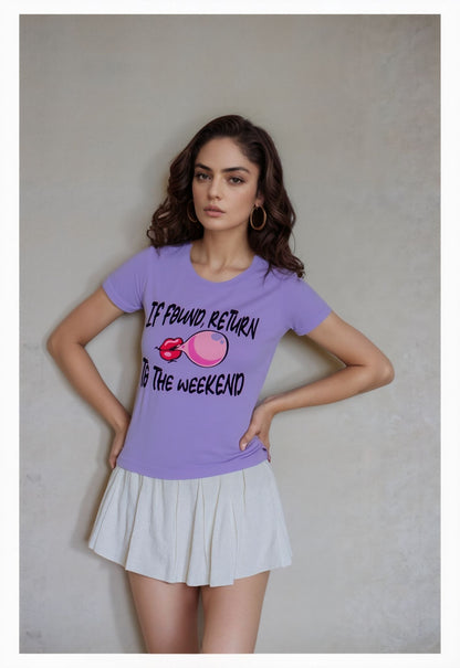 Woman wearing a lavender graphic t-shirt with the text "If Found, Return to the Weekend" and a cartoon bubble graphic, paired with a white pleated mini skirt, standing against a neutral background.


