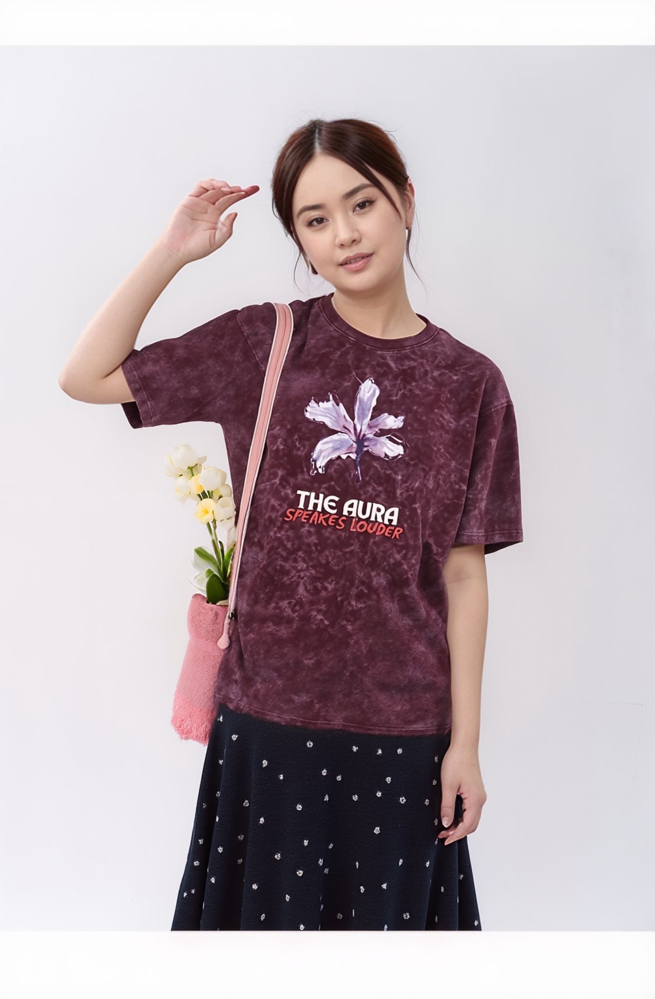 Woman wearing a maroon t-shirt with a logo and text, holding a pink bag with flowers.