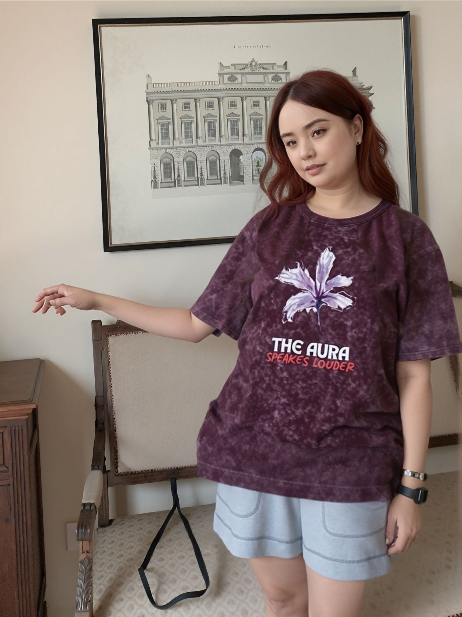 Person wearing a purple t-shirt with text and graphics, standing in a room with a framed picture on the wall.