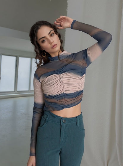 Woman posing indoors wearing a ruched mesh long-sleeve crop top in layered blue and nude shades paired with teal corduroy pants. She stands with one arm raised, resting her hand on her head, and gazes confidently at the camera.