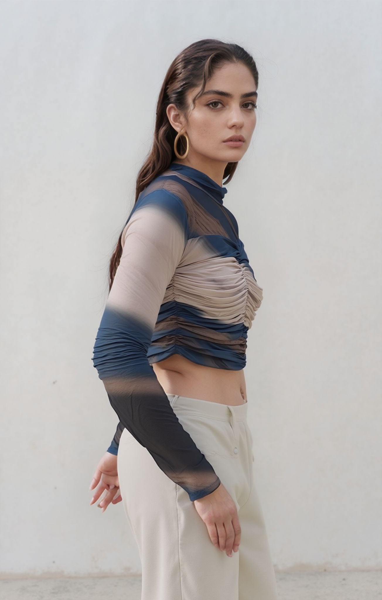 Side profile of a woman wearing a ruched mesh long-sleeve crop top in layered blue, nude, and brown tones, paired with high-waisted cream pants. She looks directly at the camera with a serious expression.