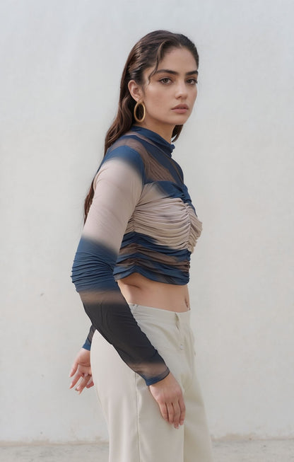 Side profile of a woman wearing a ruched mesh long-sleeve crop top in layered blue, nude, and brown tones, paired with high-waisted cream pants. She looks directly at the camera with a serious expression.