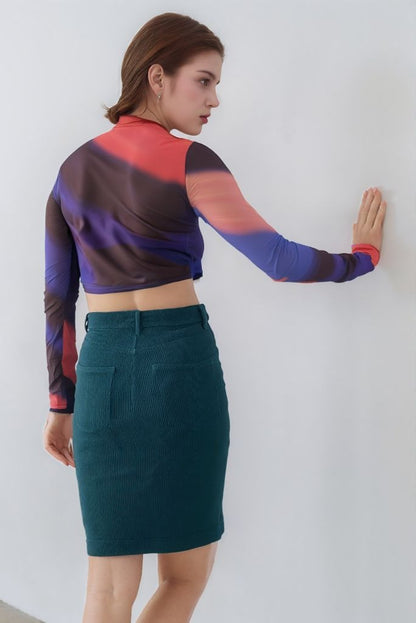 Woman wearing a colorful long-sleeve top and teal skirt against a white wall