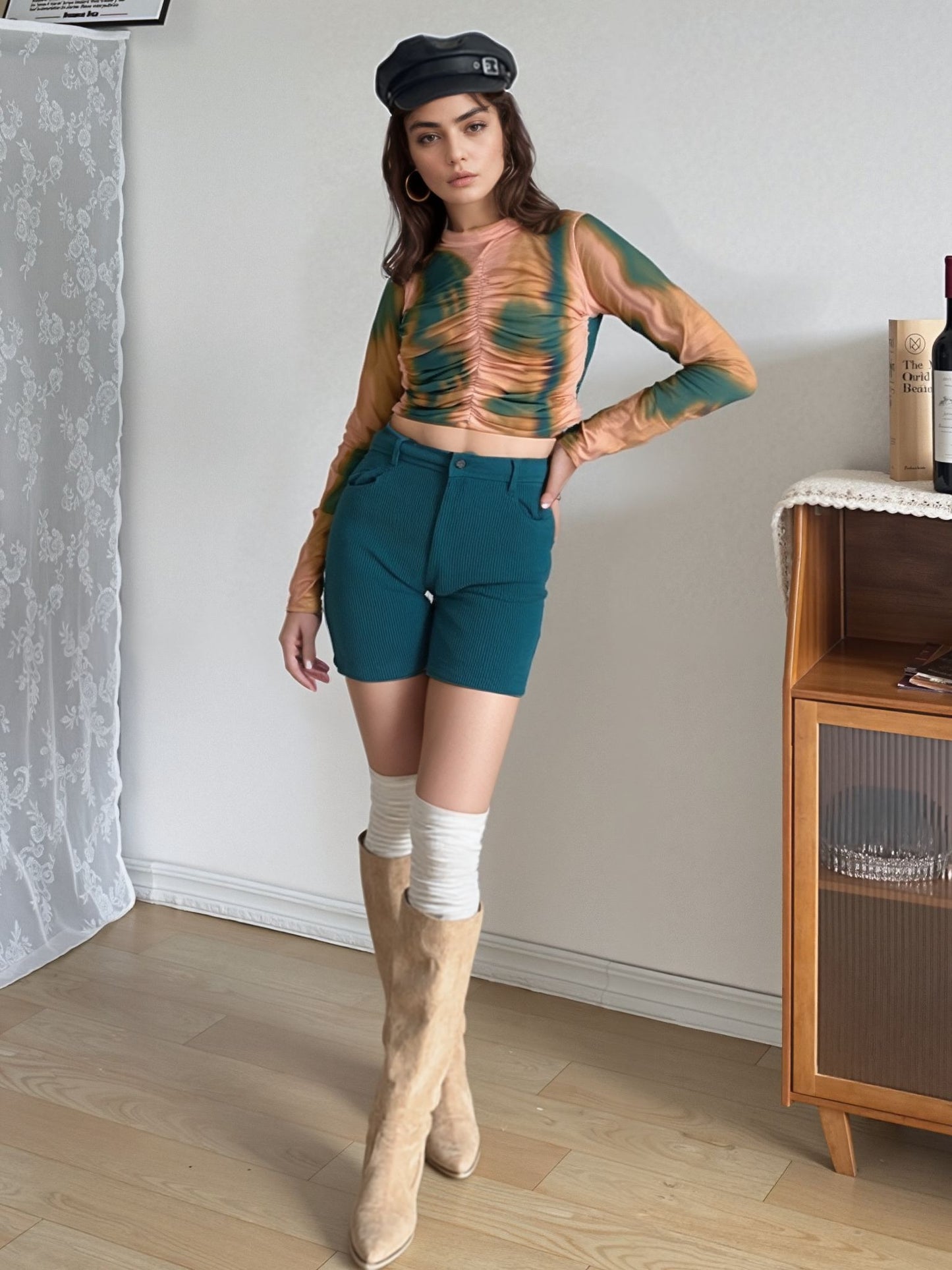 Model styled in green and mustard swirl mesh crop top with ruched front, high neckline, and full sleeves, paired with teal shorts, knee-high socks, furry boots, and black beret for a retro-inspired look.