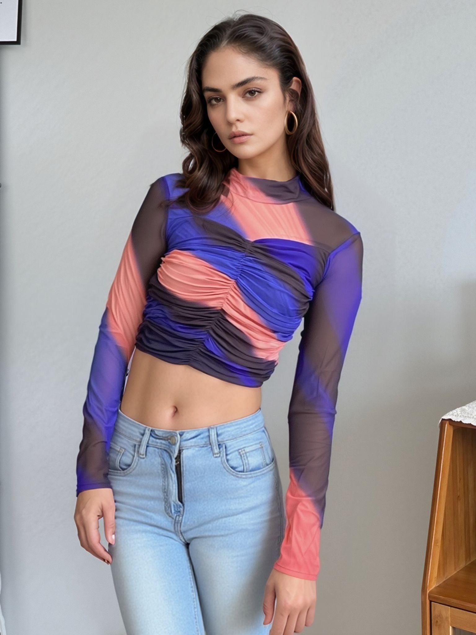 Model wearing vibrant colorblock mesh ruched crop top featuring shades of blue, coral pink, and black with sheer long sleeves and high neckline, paired with light blue high-waisted jeans