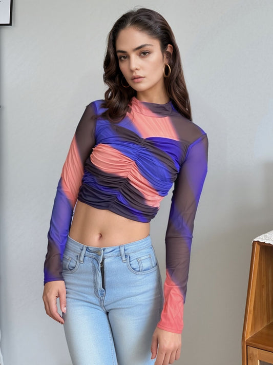 Model wearing vibrant colorblock mesh ruched crop top featuring shades of blue, coral pink, and black with sheer long sleeves and high neckline, paired with light blue high-waisted jeans