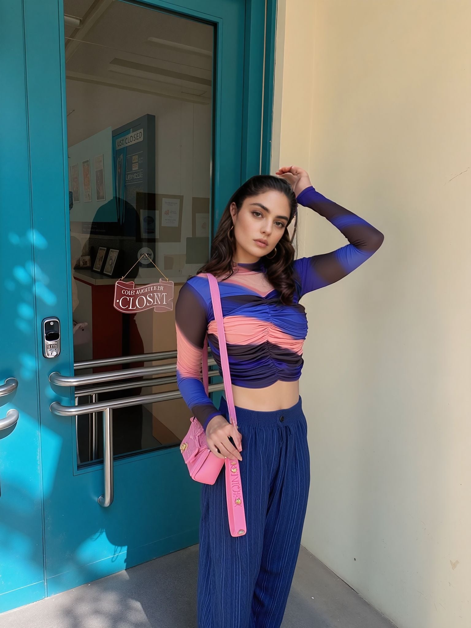 Model wearing a bold colorblock mesh ruched crop top in blue, coral pink, and black with sheer sleeves, paired with loose blue pants and a pink shoulder bag, posing outside near a teal door