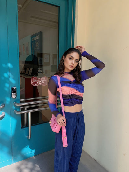 Model wearing a bold colorblock mesh ruched crop top in blue, coral pink, and black with sheer sleeves, paired with loose blue pants and a pink shoulder bag, posing outside near a teal door