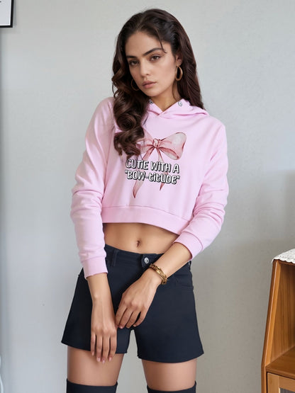 Woman wearing a pink cropped hoodie with a bow graphic and the text "Cutie with a 'Bow-titude'," paired with black shorts and hoop earrings, standing indoors.