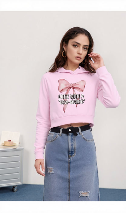 Woman wearing a pink cropped hoodie with a bow design and “Cutie with a Bow-titude” text, paired with a high-waisted ripped denim skirt.

