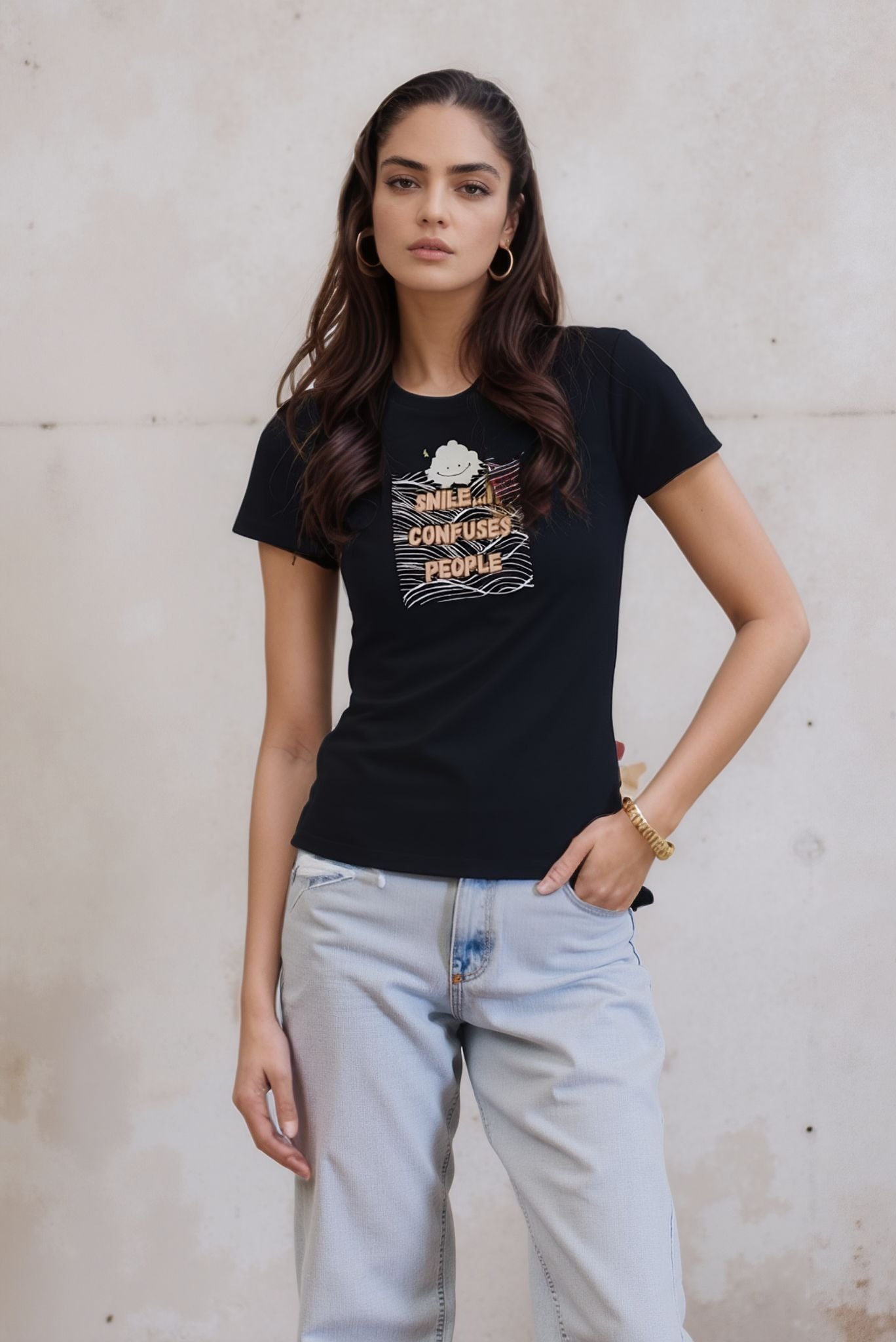 Woman modeling a black graphic t-shirt that reads "Smile. It Confuses People," styled with light-wash jeans. She stands outdoors against a textured beige wall, with one hand in her pocket.