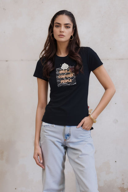 Woman modeling a black graphic t-shirt that reads "Smile. It Confuses People," styled with light-wash jeans. She stands outdoors against a textured beige wall, with one hand in her pocket.