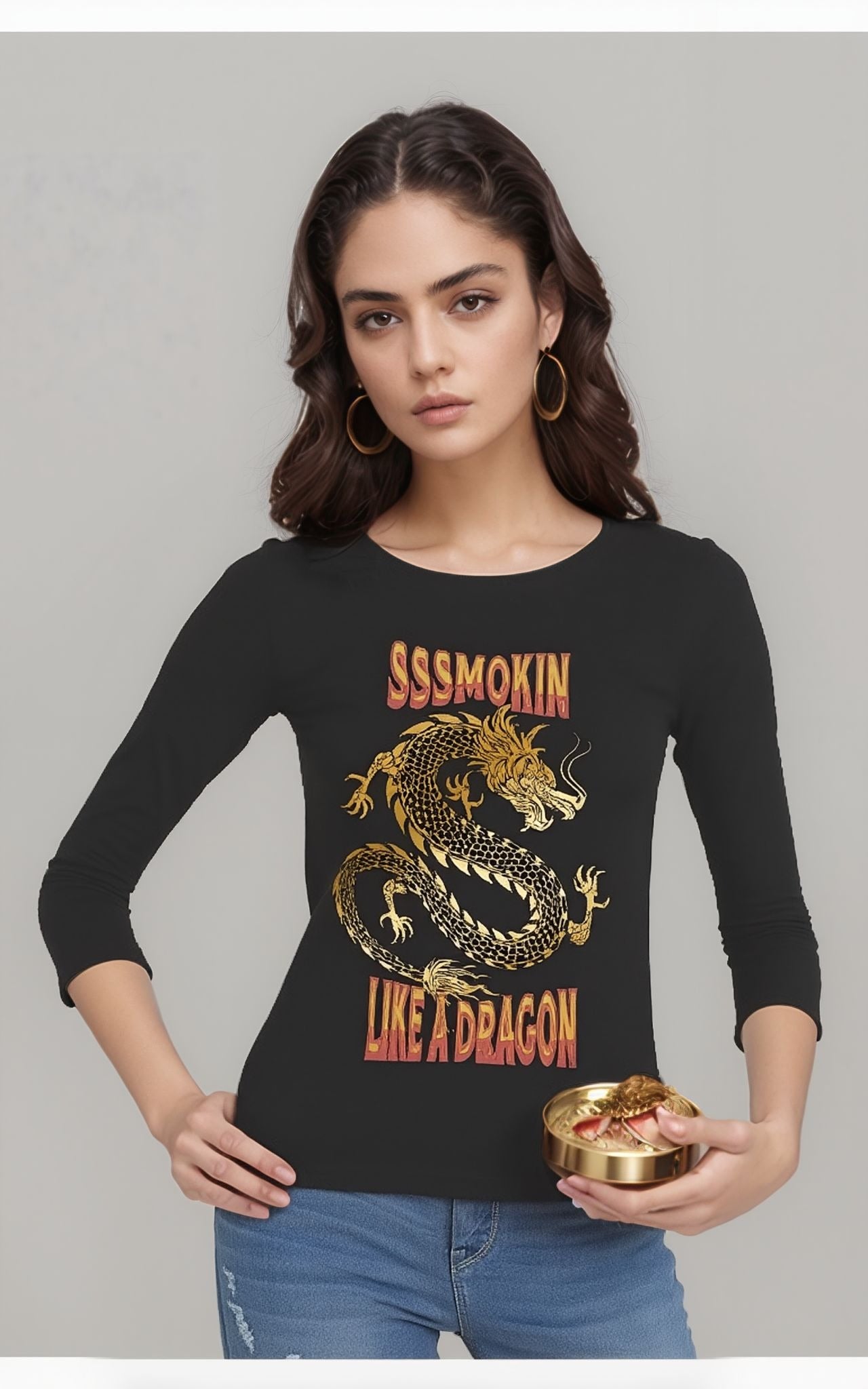 Woman wearing a black long-sleeve t-shirt featuring a bold golden dragon graphic and the text “SSSmokin Like a Dragon” in red-yellow gradient font, paired with blue jeans and gold hoop earrings, holding a round gold container.