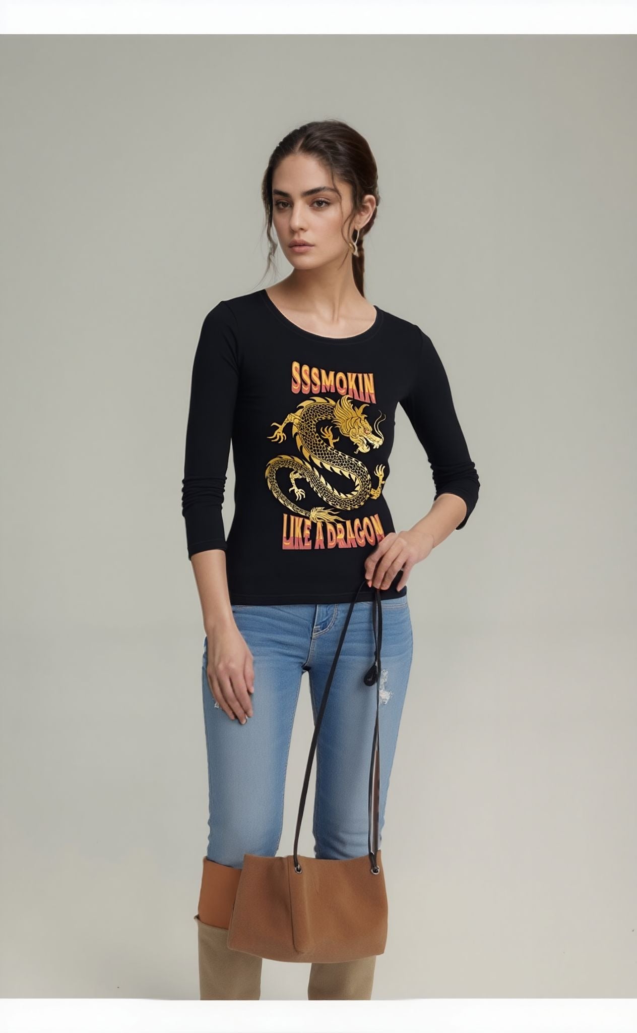 Woman wearing a black graphic t-shirt with a bold gold dragon design and the text “SSSmokin Like a Dragon,” paired with light blue ripped skinny jeans and knee-high boots, holding a tan handbag with black straps.