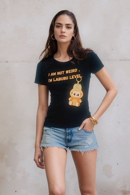 A woman wearing a black graphic t-shirt that says "I am not weird, I'm Labubu level" with a cute cartoon character illustration, paired with distressed denim shorts, standing against a plain light-colored wall.