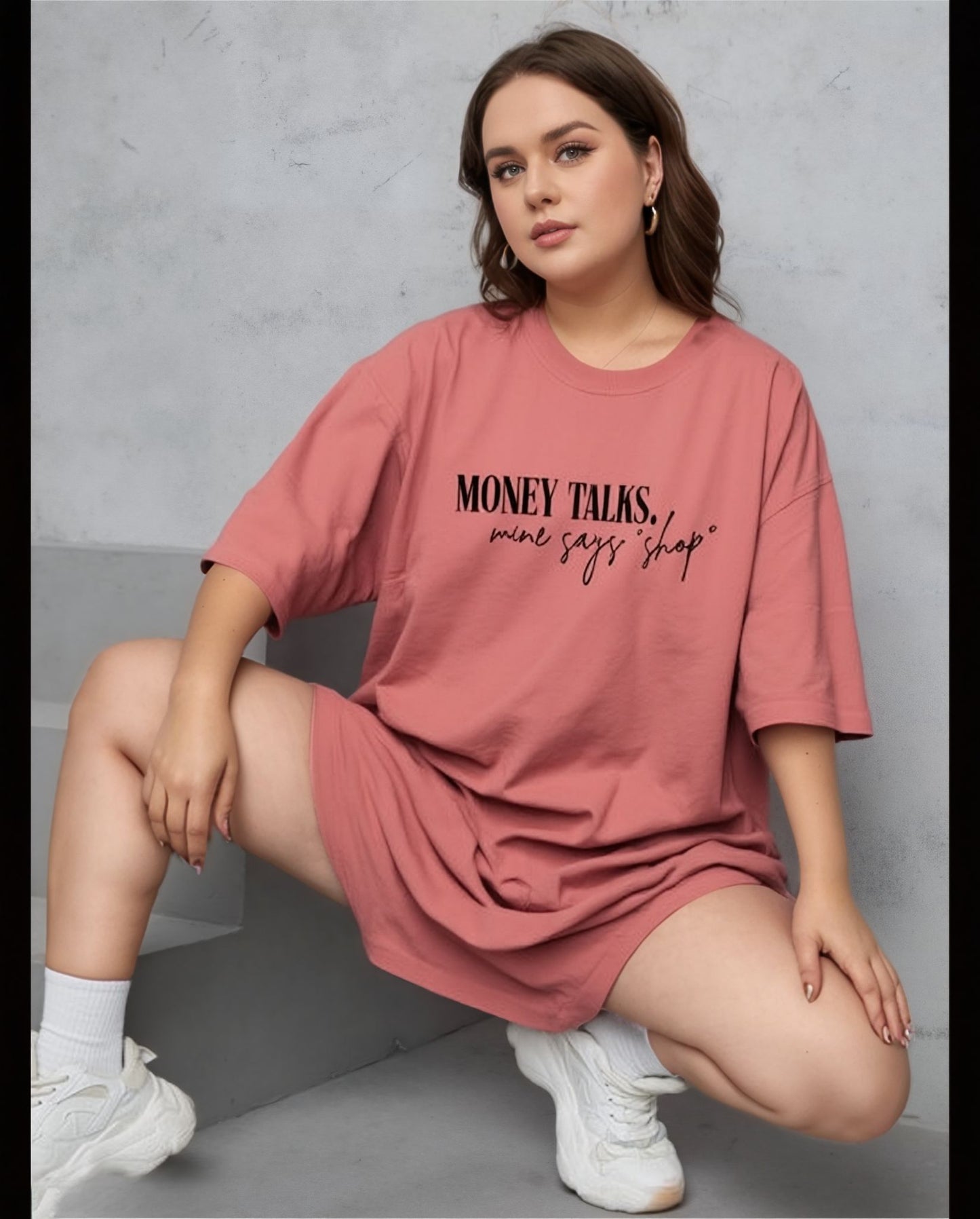 Woman wearing a pink oversized t-shirt with text, sitting on a gray surface.