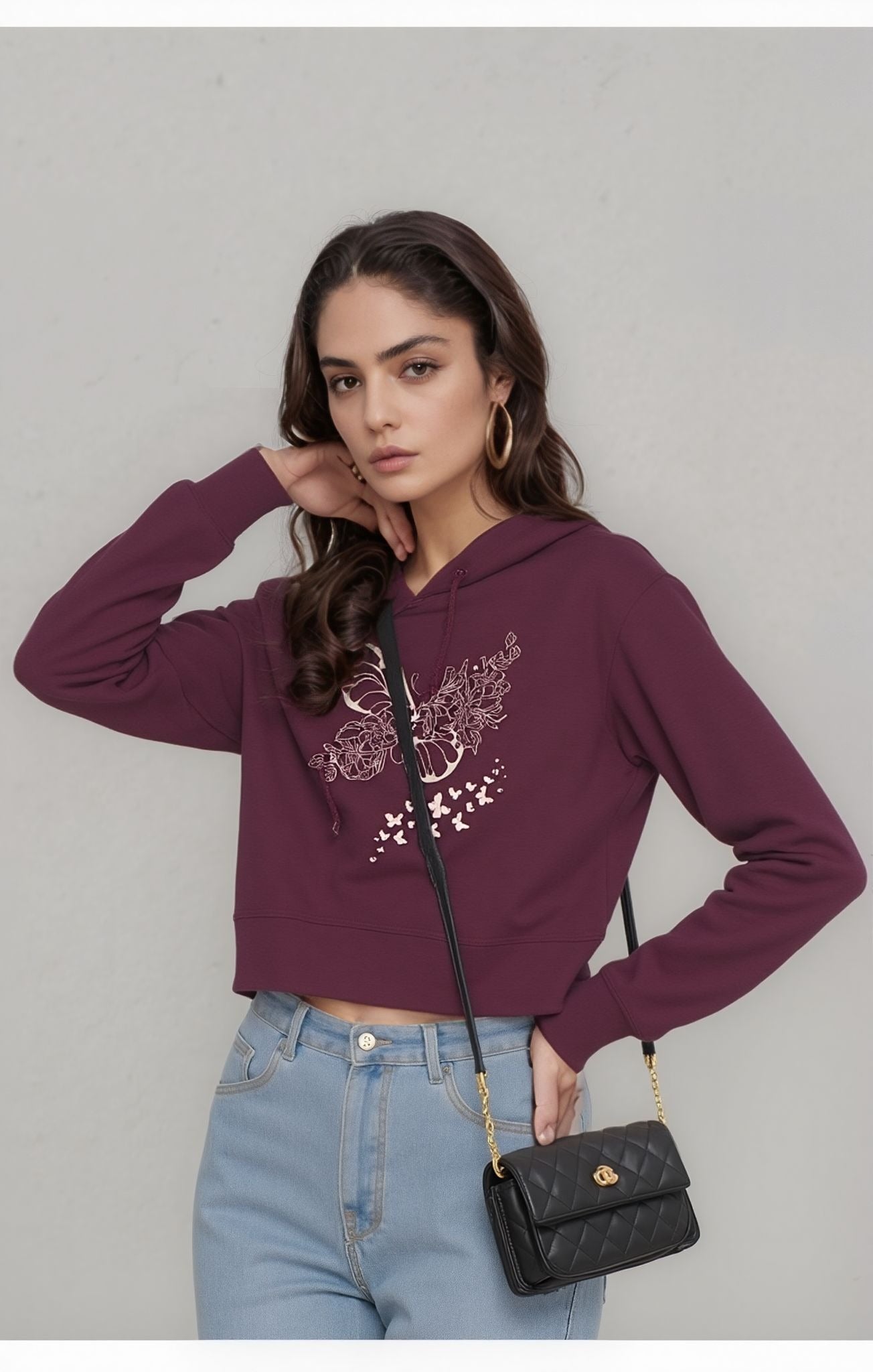 A woman wearing a maroon cropped hoodie with a floral butterfly print, light blue high-waisted jeans, and a black quilted shoulder bag, posing against a grey background.
