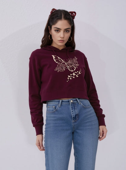 Rear view of a woman wearing a plain maroon cropped hoodie with a hood, paired with fitted light jeans, standing against a white background.