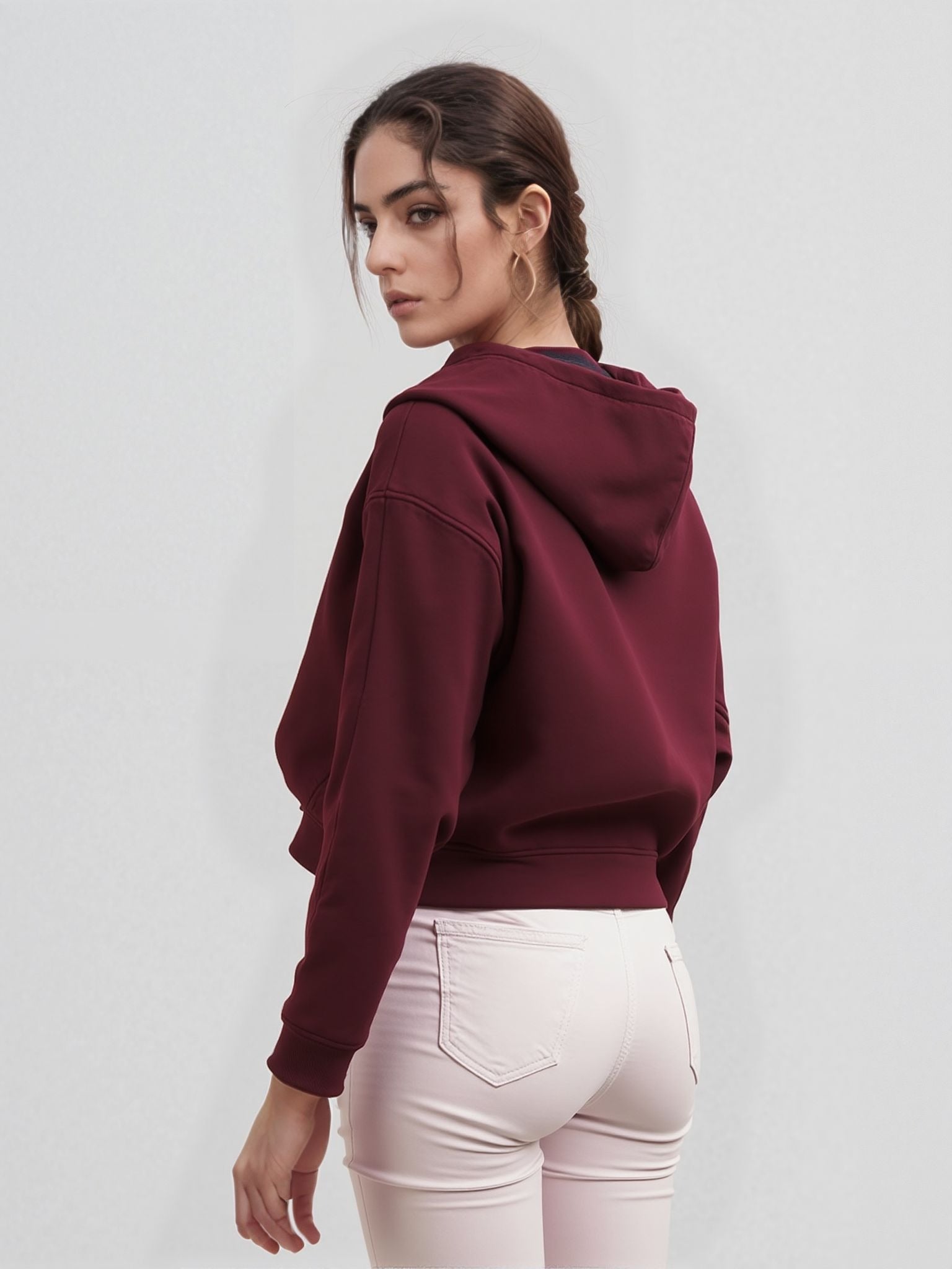 Rear view of a woman wearing a plain maroon cropped hoodie with a hood, paired with fitted light pink jeans, standing against a white background.