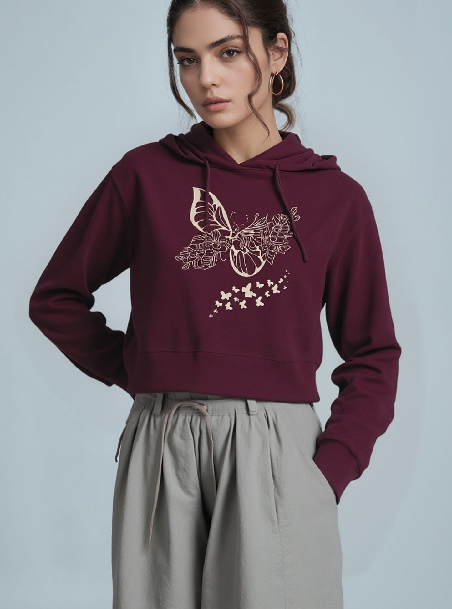 A woman wearing a maroon cropped hoodie with a beige floral and butterfly graphic on the front, paired with light grey high-waisted drawstring pants, standing against a light blue background.