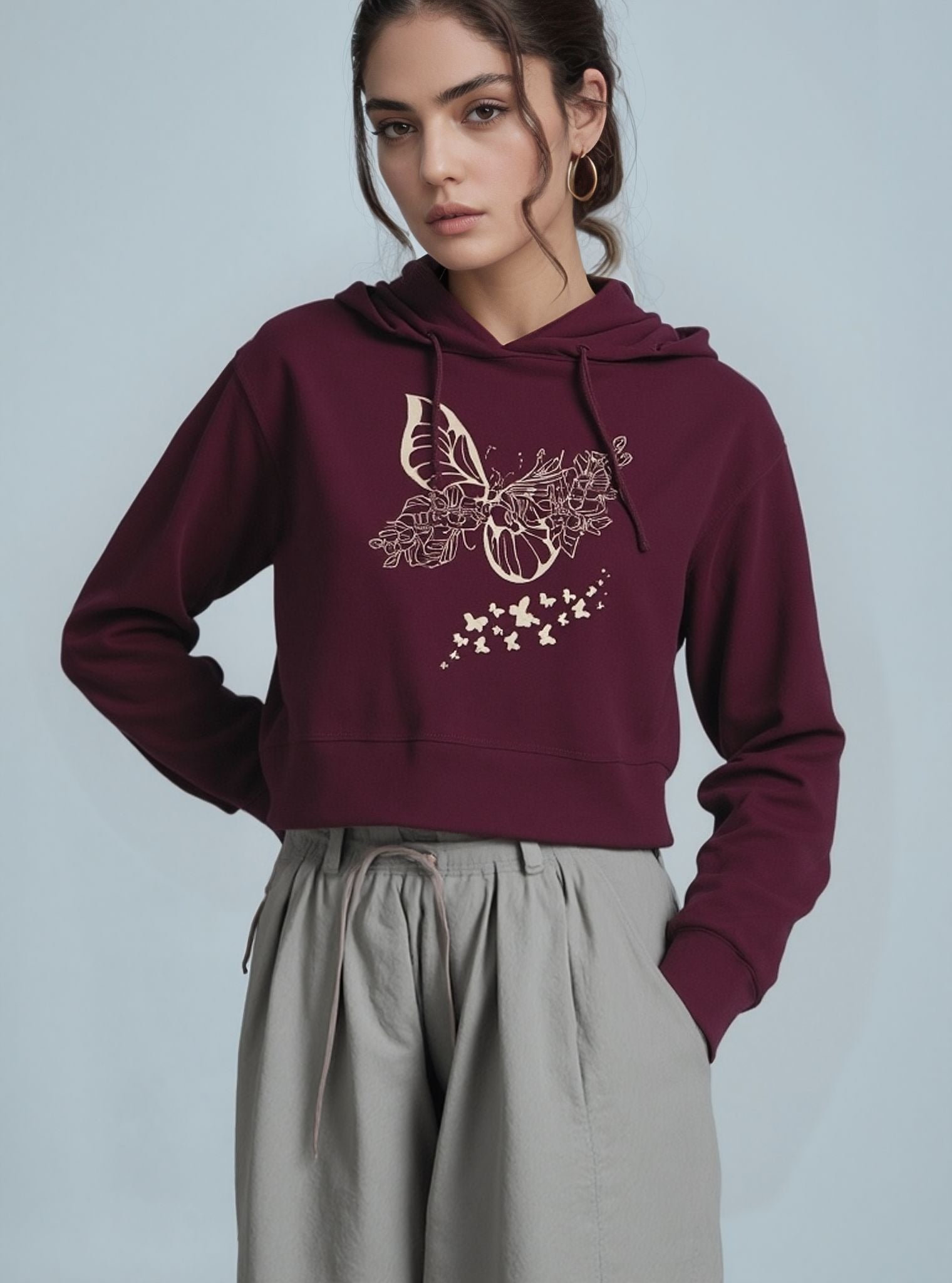 A woman wearing a maroon cropped hoodie with a beige floral and butterfly graphic on the front, paired with light grey high-waisted drawstring pants, standing against a light blue background.