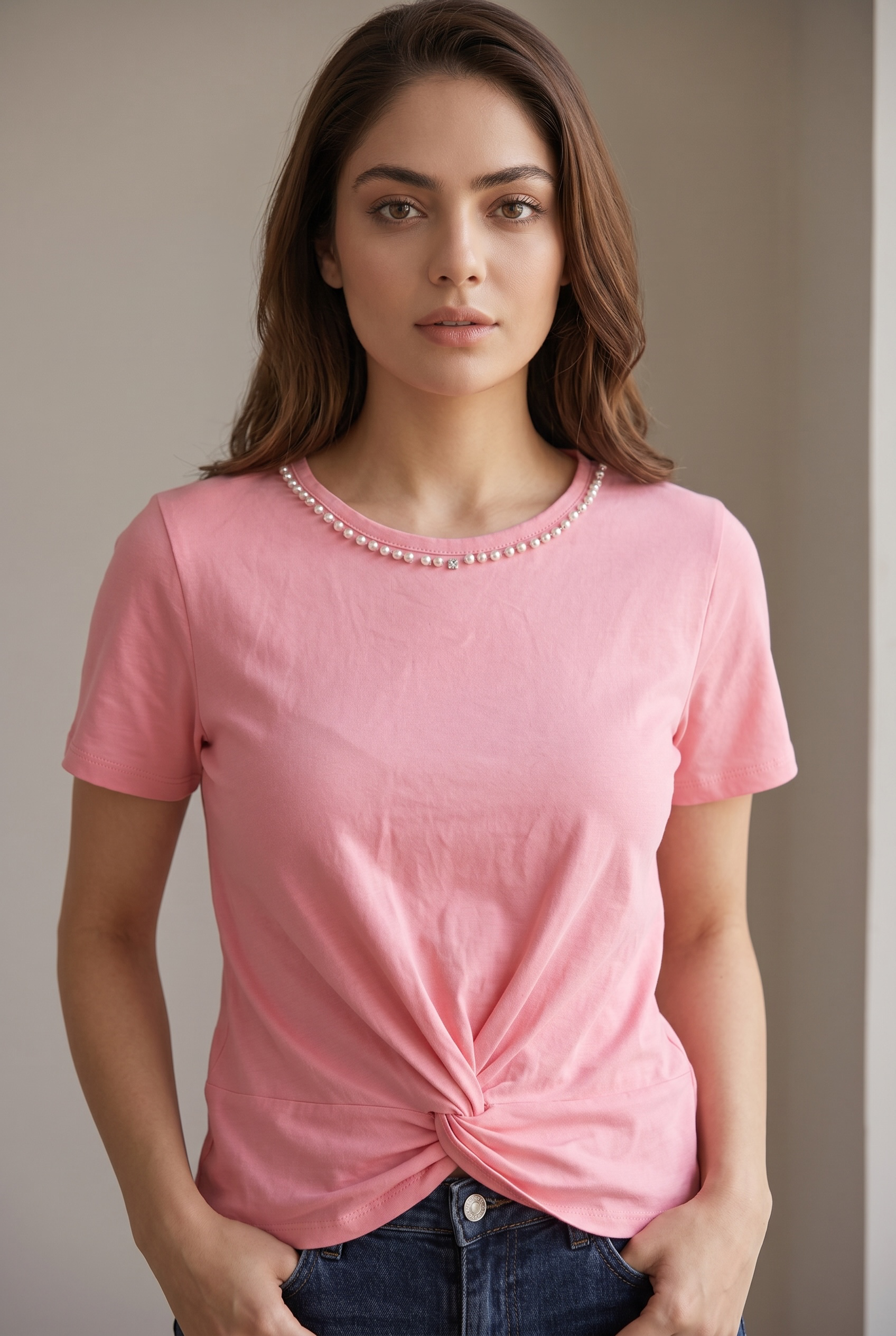 Front portrait pose of a woman wearing a blush pink pearl embellished twist top paired with dark blue denim and hands in pockets.