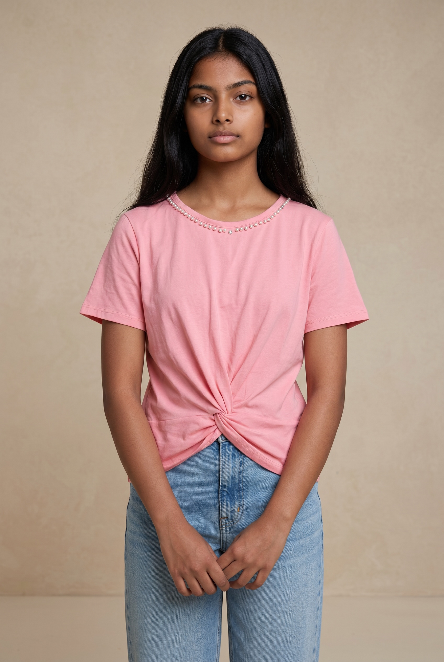 Front standing pose of a woman wearing a blush pink cotton top with pearl neckline and front twist detail in natural light.
