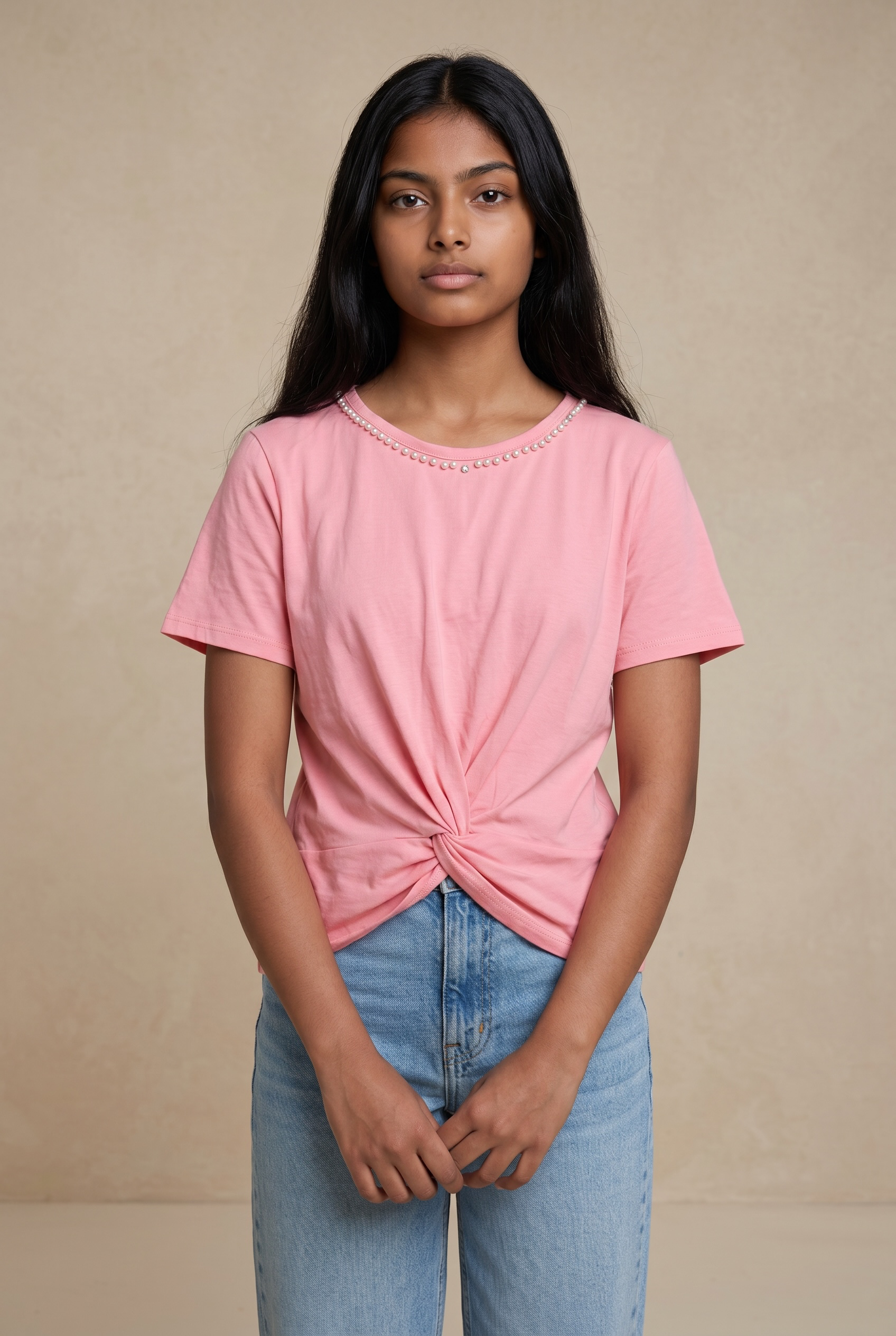 Front standing pose of a woman wearing a blush pink cotton top with pearl neckline and front twist detail in natural light.
