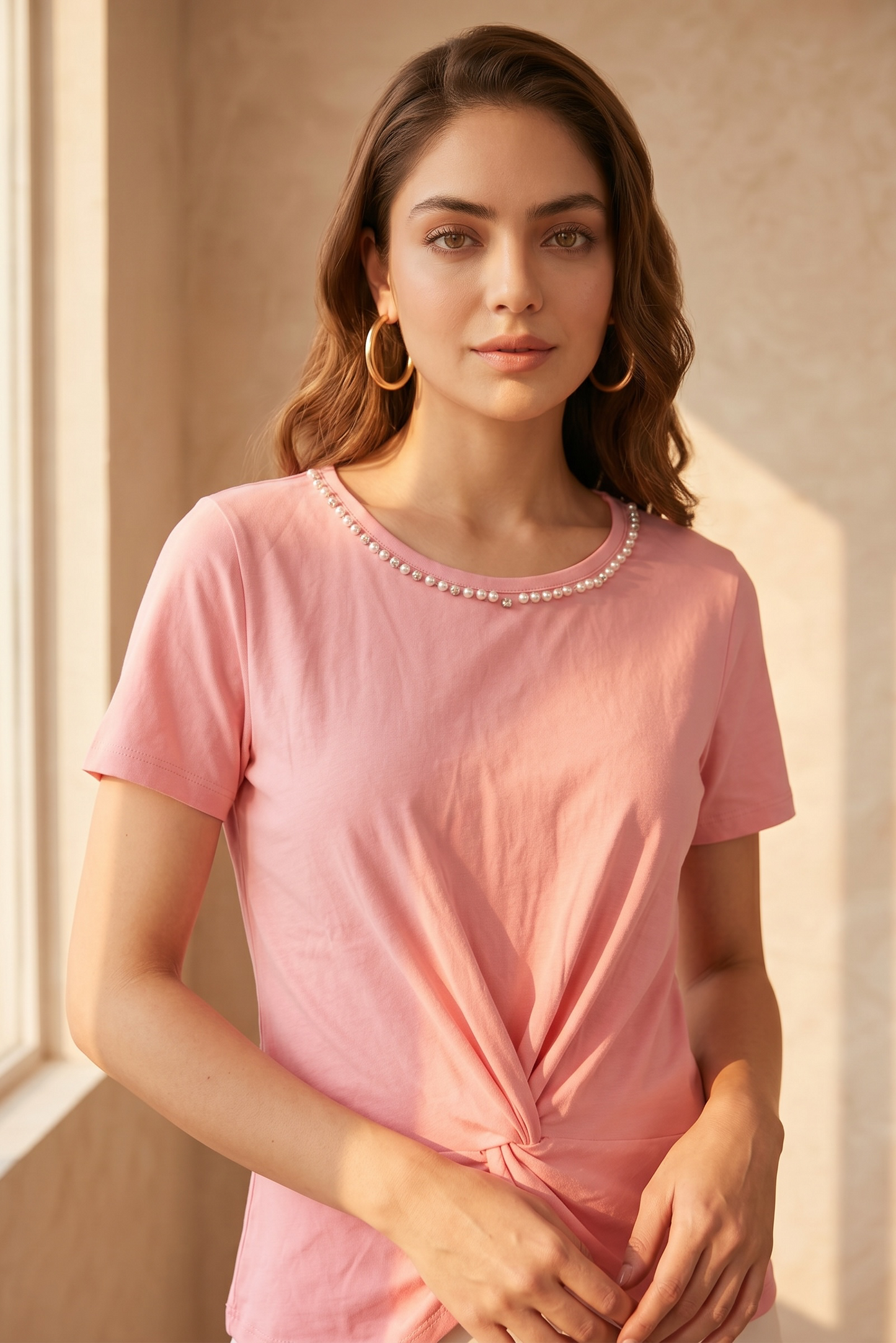Front standing portrait pose of a woman wearing a blush pink pearl neck twist top beside a window in natural light.
