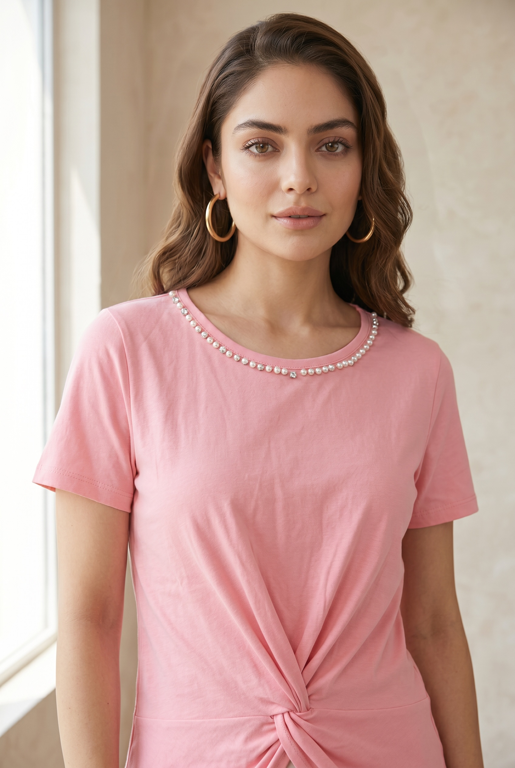 Front standing pose of a model wearing a blush pink pearl neck twist top paired with light blue jeans against a neutral studio background.
