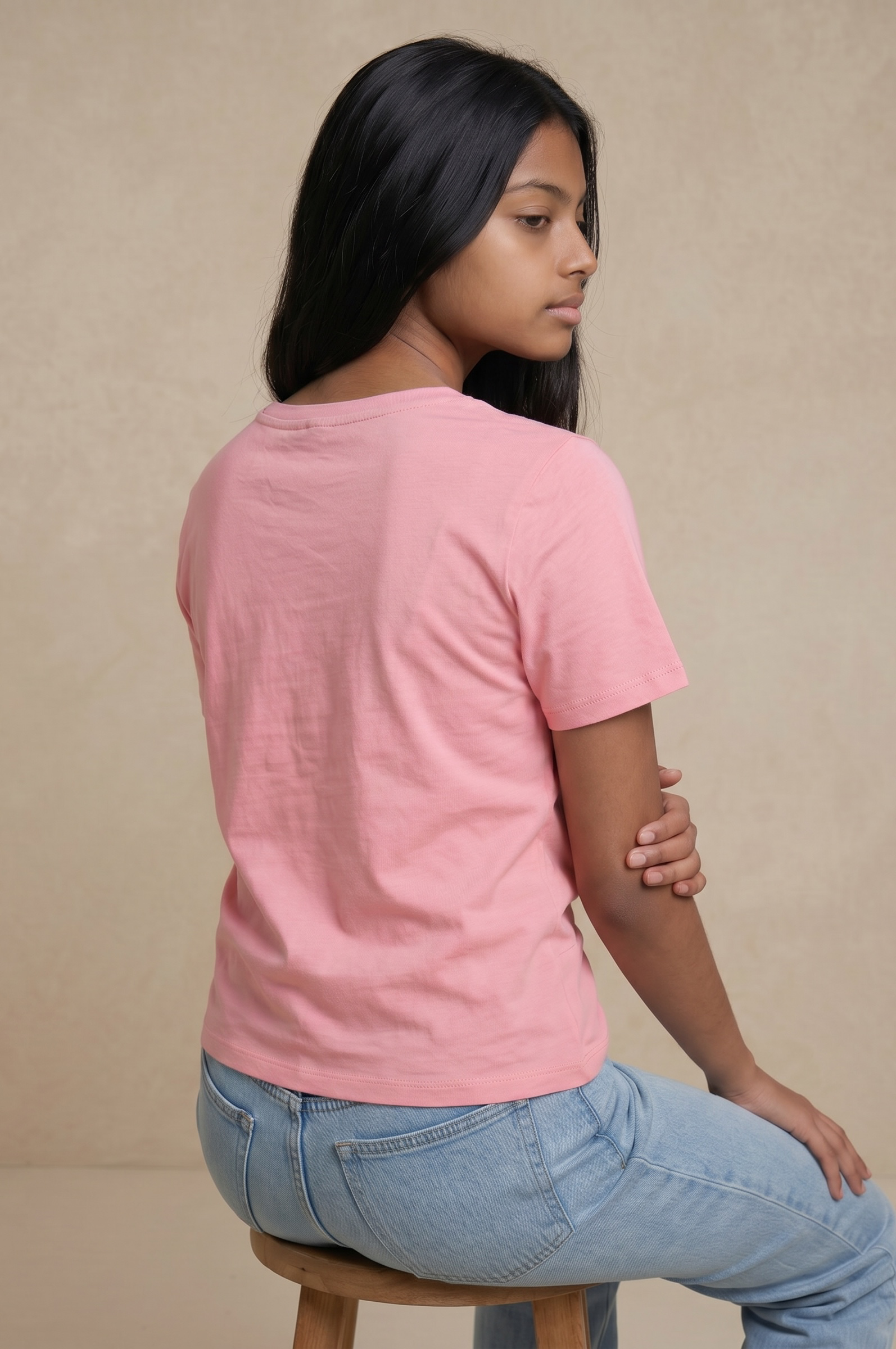 Back sitting pose of a woman wearing a blush pink cotton twist top with a pearl embellished neckline and light blue jeans.
