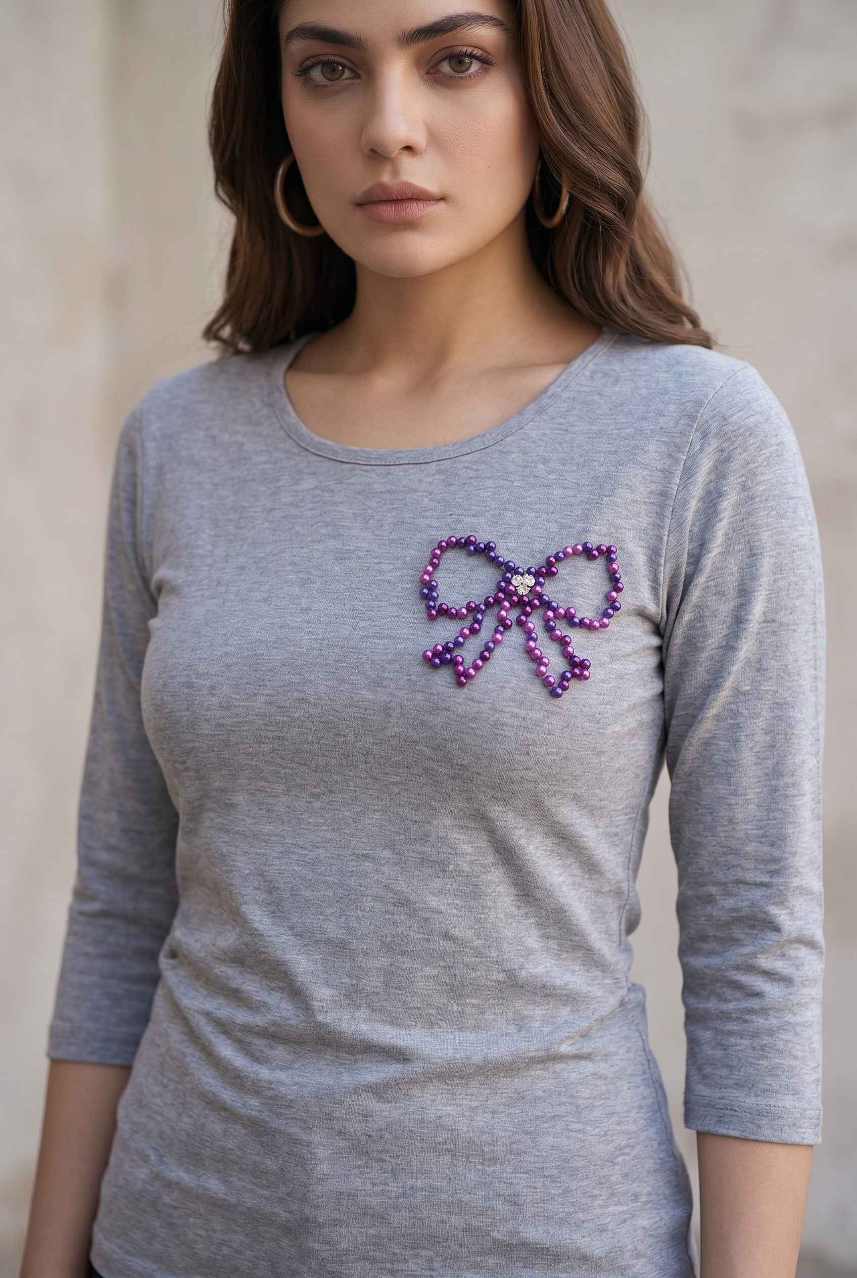 Woman wearing a grey full sleeve cotton T-shirt featuring a hand-embellished purple pearl bow design.
