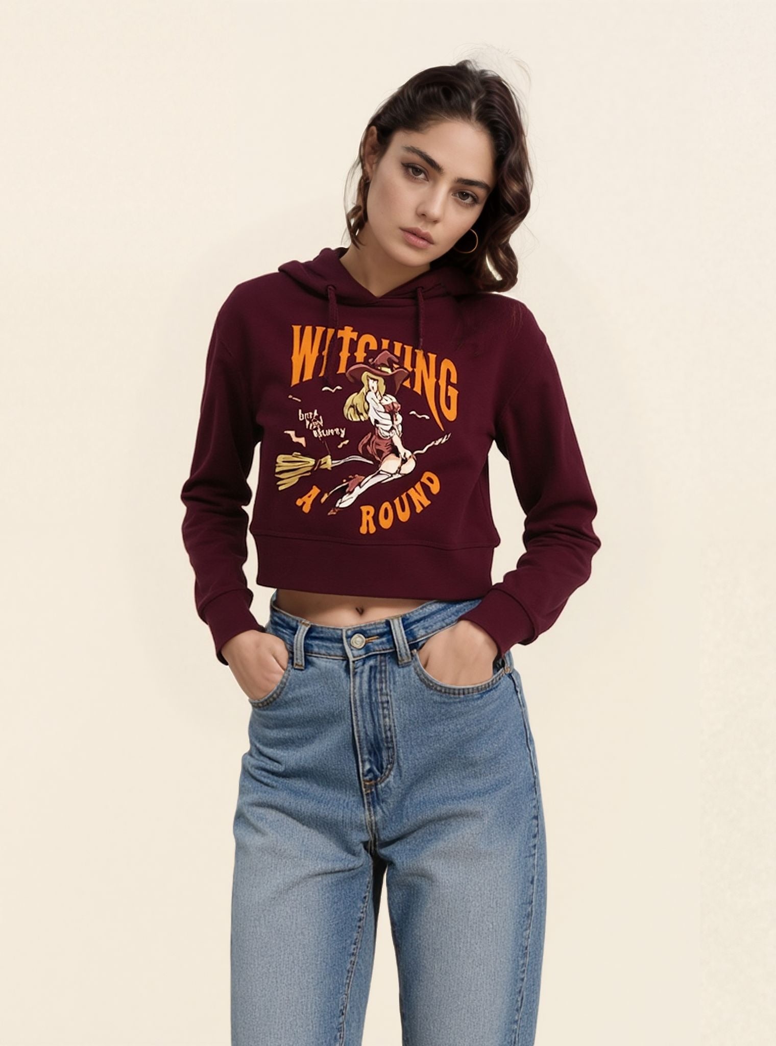 A woman wearing a maroon cropped hoodie with 'WITCHING AROUND' graphic in orange and white, paired with blue jeans.