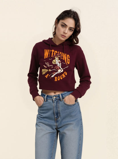 A woman wearing a maroon cropped hoodie with 'WITCHING AROUND' graphic in orange and white, paired with blue jeans.