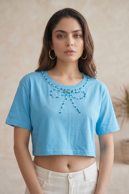Front-facing portrait pose of a woman wearing a sky blue cotton crop top with a handcrafted pearl bow neckline.
