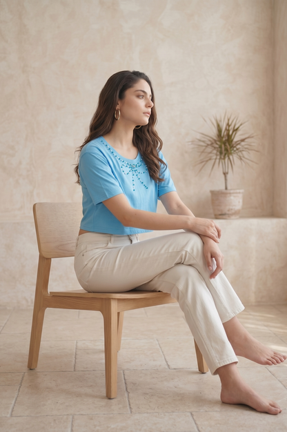Side sitting pose of a woman on a wooden chair wearing a sky blue pearl bow crop top paired with cream trousers.

