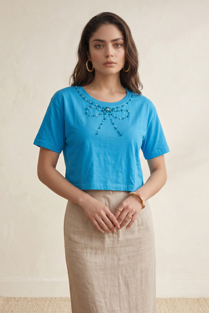 Front standing pose of a woman wearing a sky blue crop top featuring a handcrafted pearl bow neckline paired with a beige skirt.