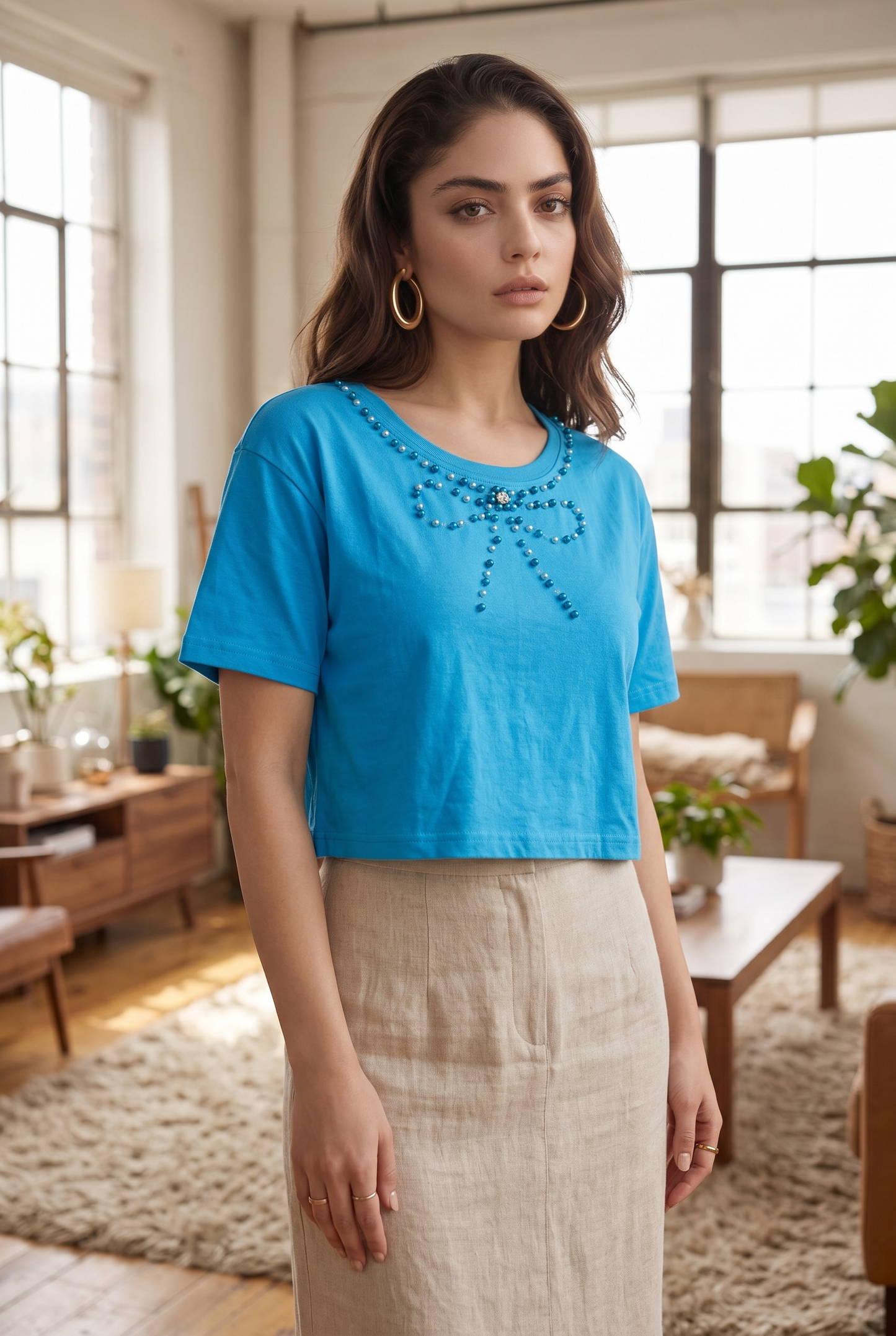 Standing three-quarter pose of a woman wearing a sky blue cotton crop top with pearl bow embellishment in a modern living room.
